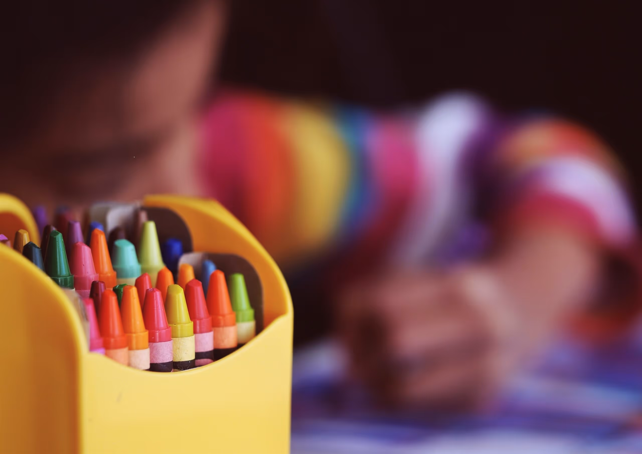 Close up of brightly coloured crayons whilst a child colours in soft focus in the background.