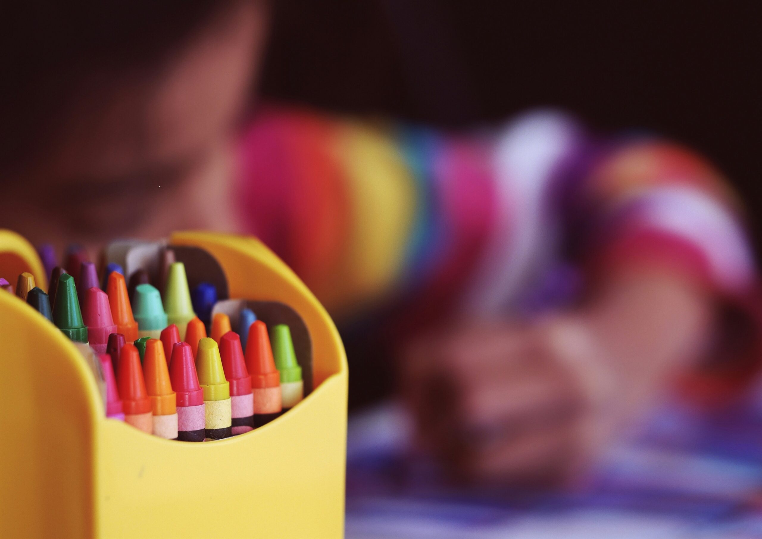 Close up of brightly coloured crayons whilst a child colours in soft focus in the background.