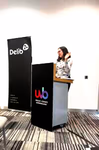 A young woman with shoulder-length dark brown hair wearing a glasses and a grey jumper (Ellen Taplin from Surrey County Council) stands behind a podium and addresses an audience (who are not pictured). She is gesturing casually with her left hand.