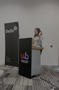 A young woman with shoulder-length dark brown hair wearing a glasses and a grey jumper (Ellen Taplin from Surrey County Council) stands behind a podium and addresses an audience (who are not pictured). She is gesturing casually with her left hand.
