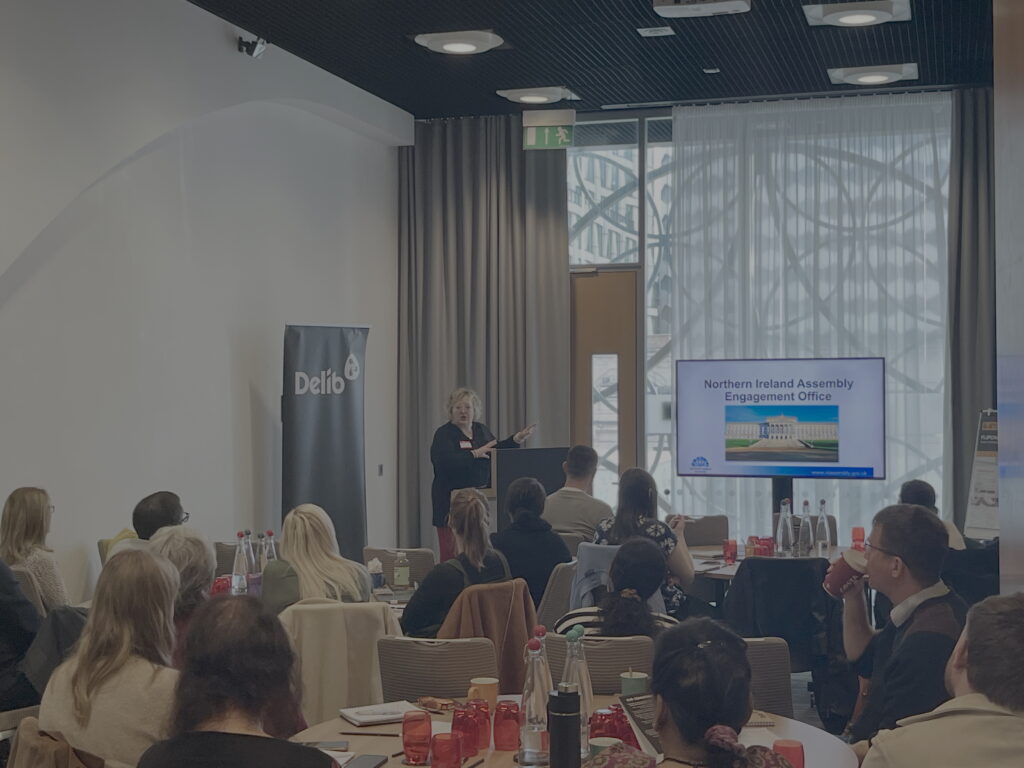 A woman with blonde hair and dressed in black (Kate McCullough from the Northern Ireland Assembly) stands beside a podium and gestures towards a large television screen to her left. She is in front of a seated audience who listen attentively to the her presentation.