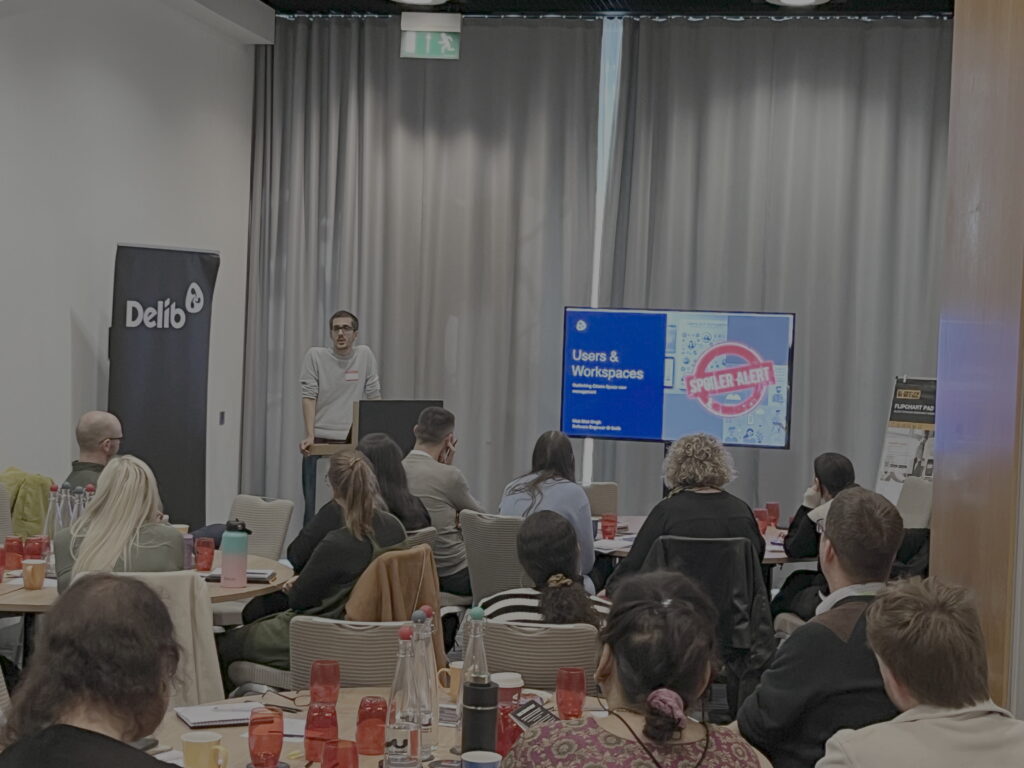 A young man with short dark hair and glasses, wearing a light grey jumper (Matt Sital-Singh from Delib) stand behind a podium and next to a large television screen in front of a seated audience who listen attentively to his presentation.