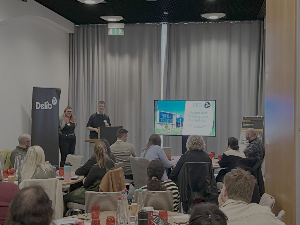 A young woman and a man (Catherine Murrell and Martin Guest from Melton Borough Council) stand behind a podium and next to a large television screen in front of a seated audience who listen attentively to their presentation. The woman has a long pinkish-red hair and wears glasses. The man has short dark hair and is also wearing glasses.