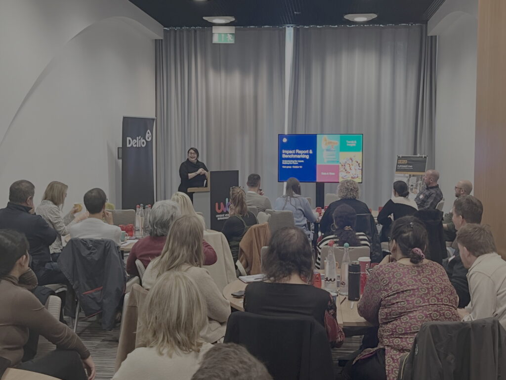 A young woman with dark hair and dressed in black (Tiffany Maddox from Delib) stands behind a podium and next to a large television screen in front of a seated audience who listen attentively to the woman's presentation. The woman smiles warmly towards the audience and has her hands clasped in front of her.
