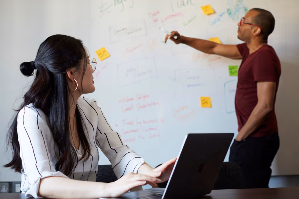 man at whiteboard with woman on computer looking as they discuss together