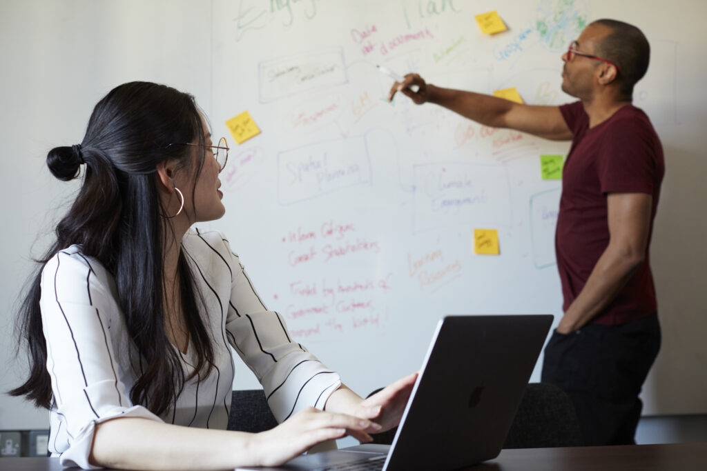 man at whiteboard with woman on computer looking as they discuss together 