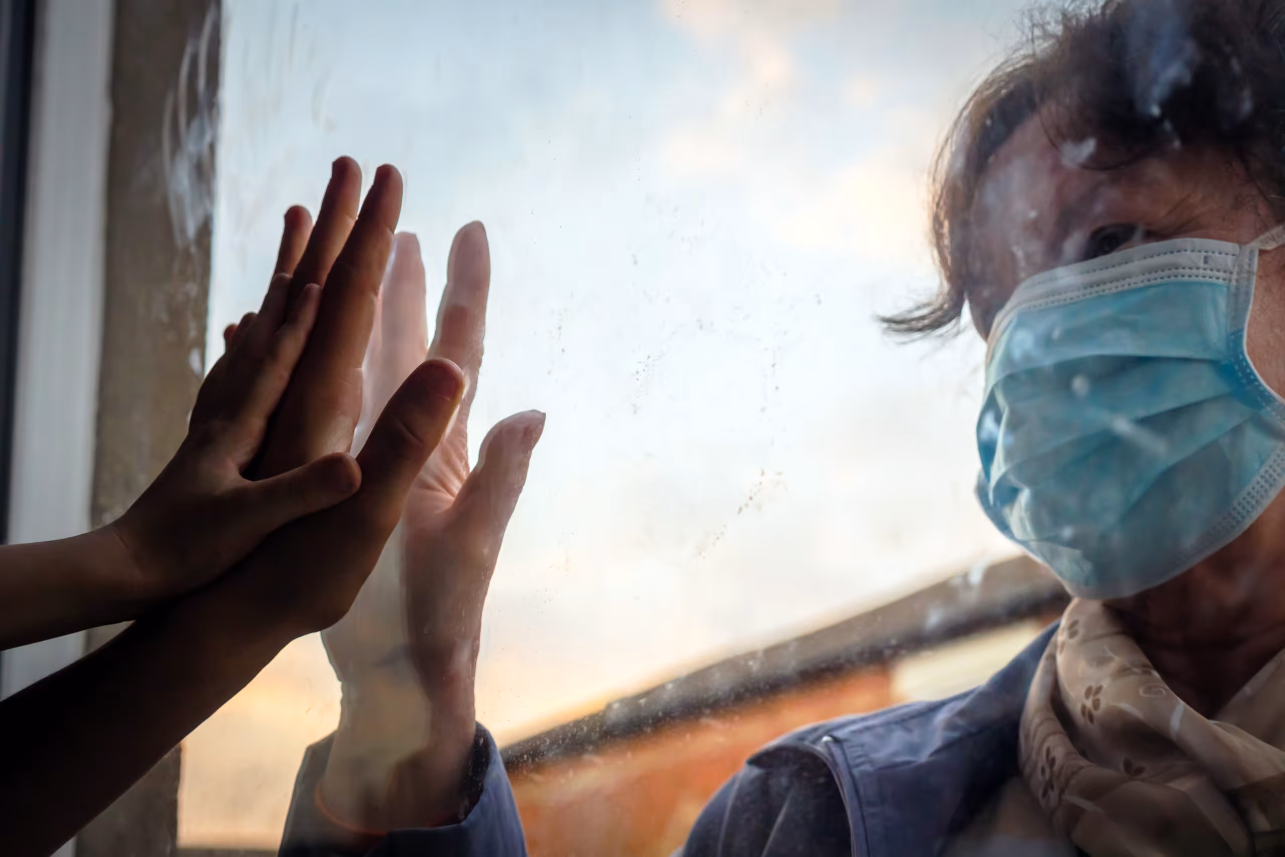 woman touches hands through glass with relative who is standing behind a window and wearing a mask