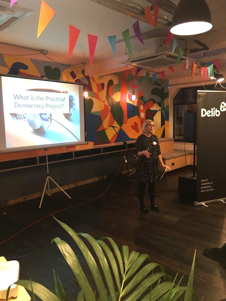 A woman standing in a brightly coloured room with a slide on a projector screen that shows the opening statement "What is the Practical Democracy Project?"