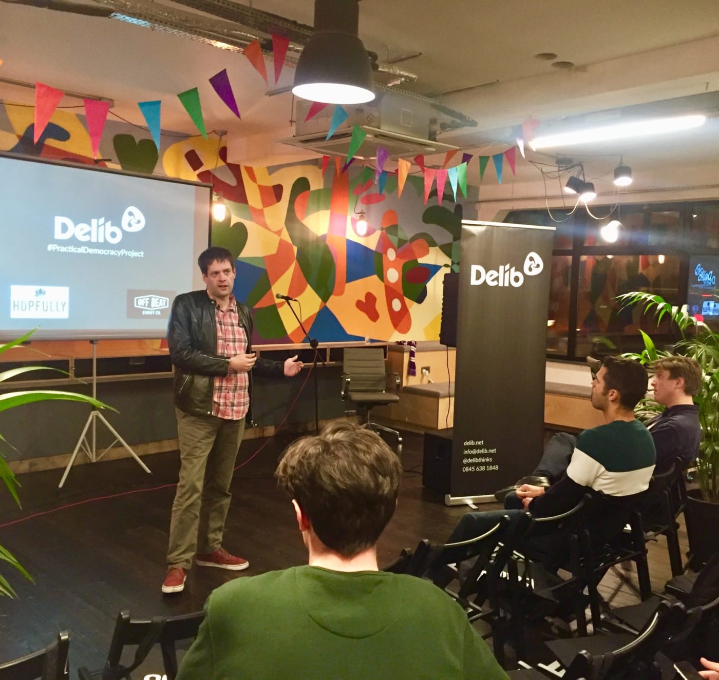Another speaker stands in front of the same slide in a brightly coloured room where you can see multiple chairs and people sat listening