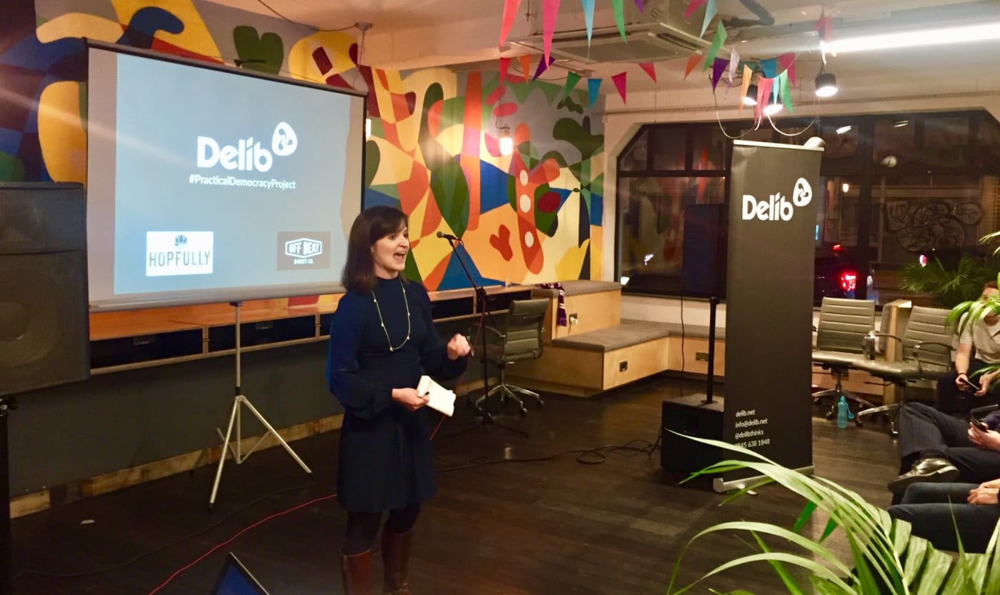 A woman standing in a brightly coloured room with a slide on a projector screen that shows a new slide with Delib's logo front and center