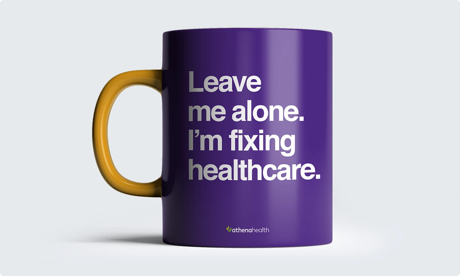 A purple athenahealth branded coffee mug designed for employees. It has a yellow handle and says "Leave me alone. I'm fixing healthcare."