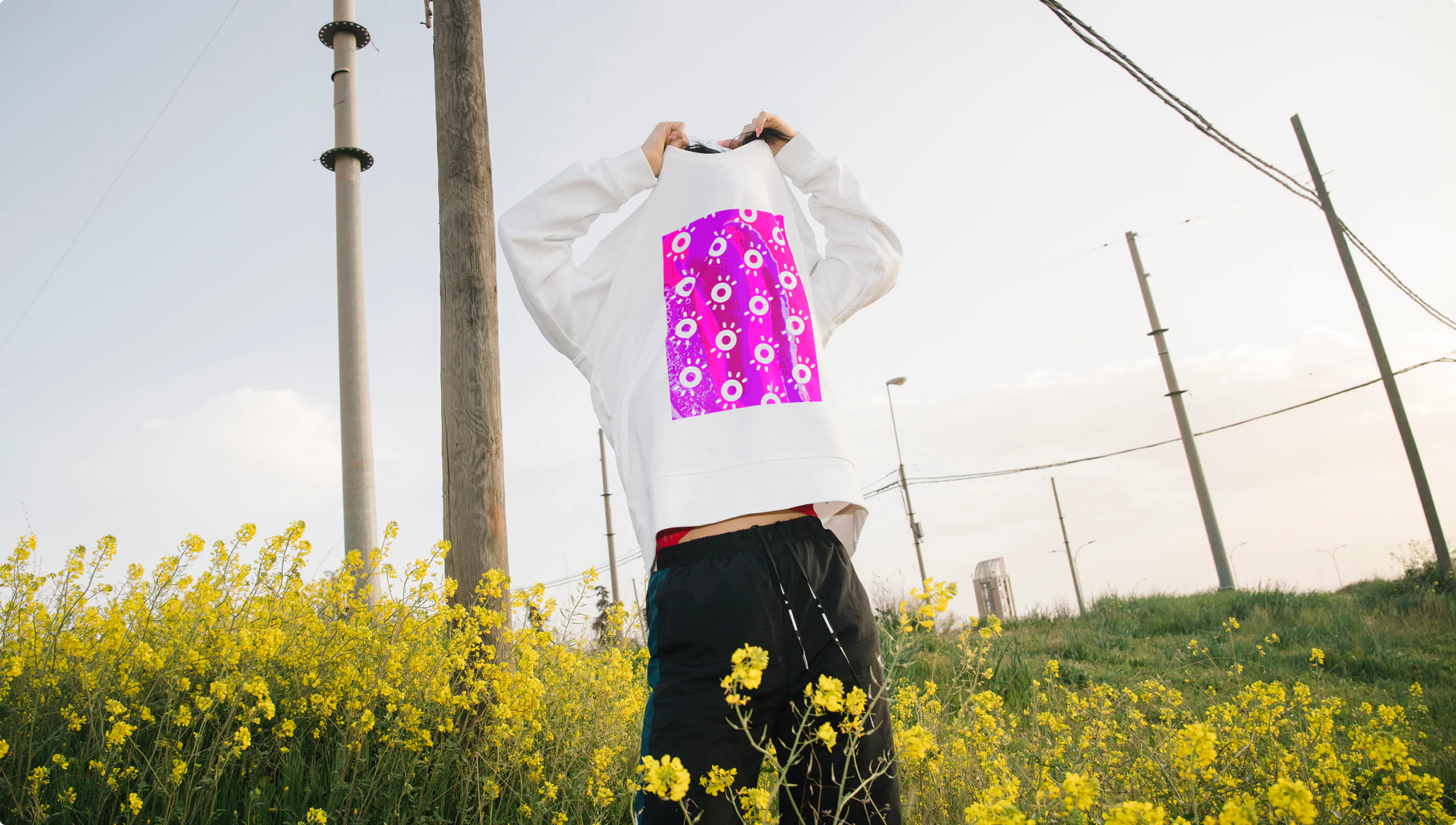 A young man removing a long sleeve white tshirt covered in Goood Wellness logos. He is standing in a field of yellow flowers.