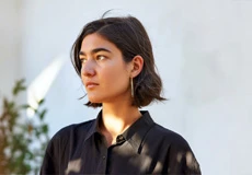Young woman with short dark hair wearing a black shirt and hoop earrings, looking to the side in natural sunlight with a white background.