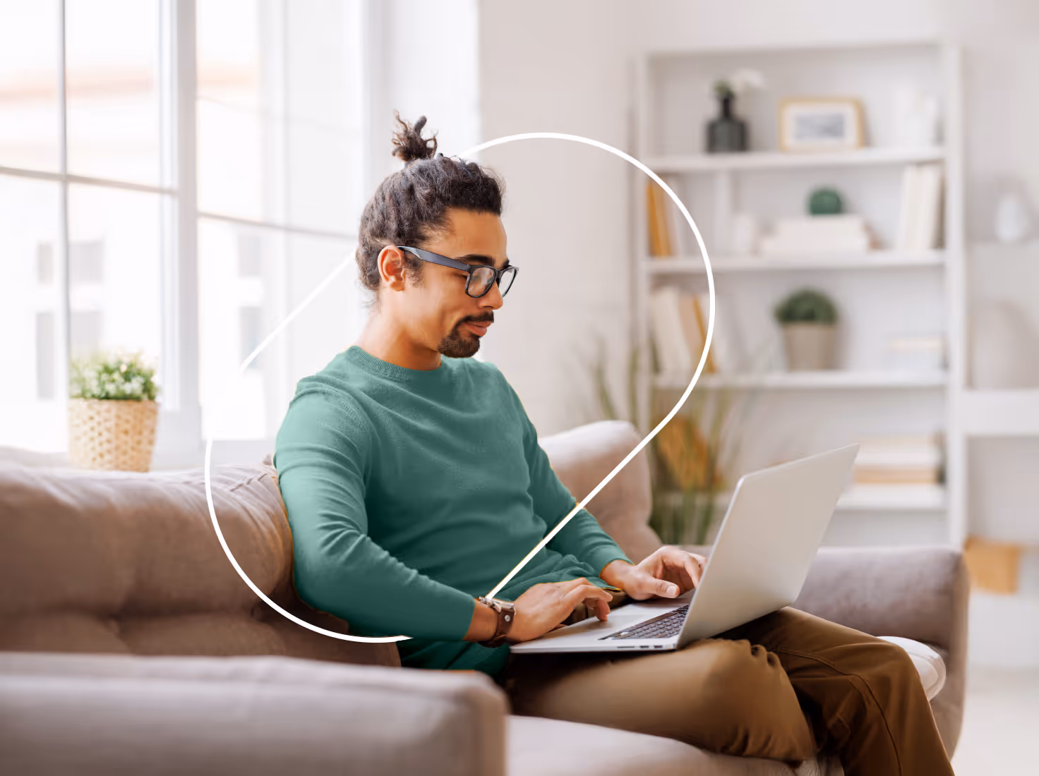 Man with glasses and a top knot working on a laptop while sitting on a couch in a bright, modern living room.