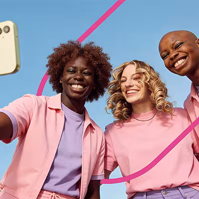 Three smiling friends in pastel-colored clothes taking a selfie against a clear blue sky.