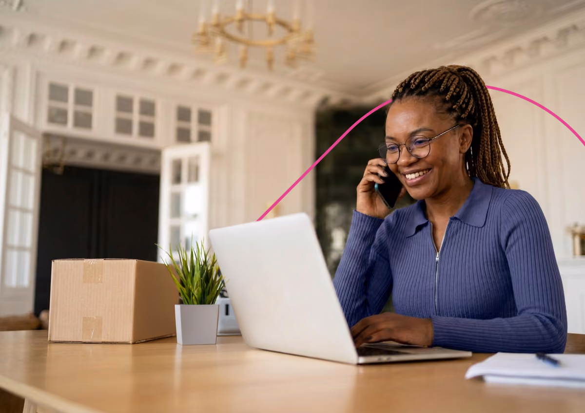 Smiling woman with braided hair working on a laptop and talking on the phone at a wooden table.