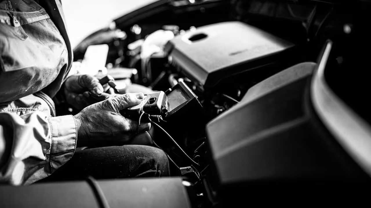 Technician registering new car battery using diagnostic tool in engine bay, close-up of tool and hands in black and white