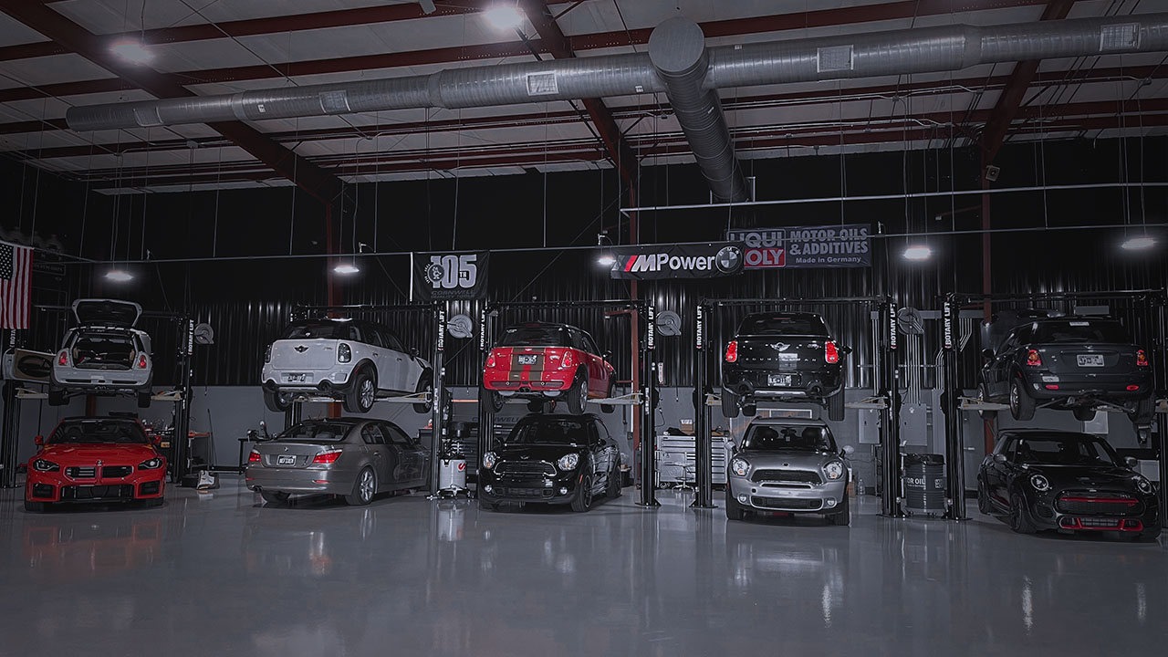 Wide-angle view of the Cooper’s Garage service bay filled with MINI Coopers on lifts, under bright shop lighting with visible Liqui Moly signage and banners overhead.