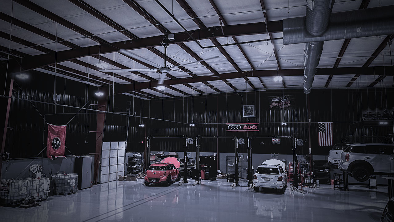 Interior of Cooper’s Garage service bay with multiple European vehicles on lifts, featuring Audi and Liqui Moly signage, Tennessee and American flags, and a polished shop floor.