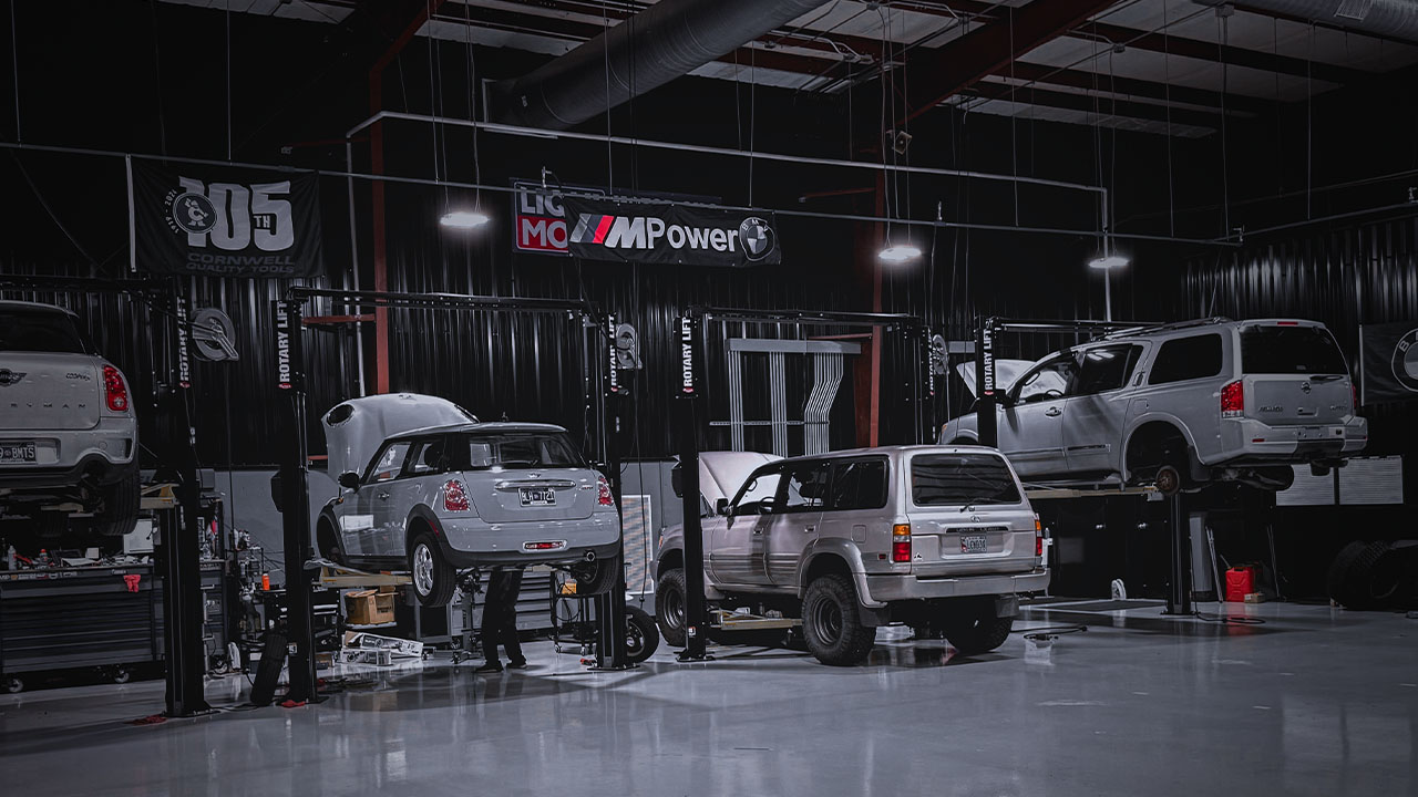 Technicians working inside Cooper’s Garage with MINI Coopers and SUVs on lifts, under MPower and Liqui Moly banners in a clean, well-lit service bay.

