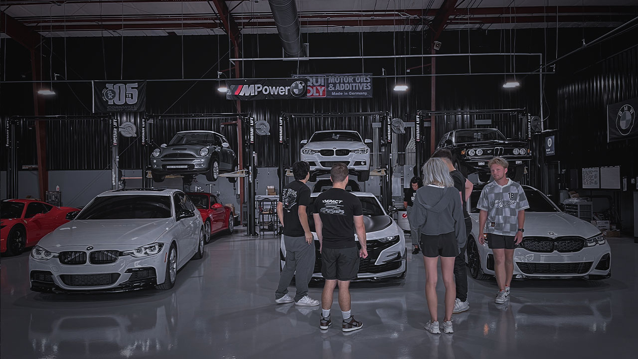 Group of visitors gathered inside the Cooper’s Garage service bay during a Cars & Coffee event, surrounded by BMWs and performance vehicles on lifts.

