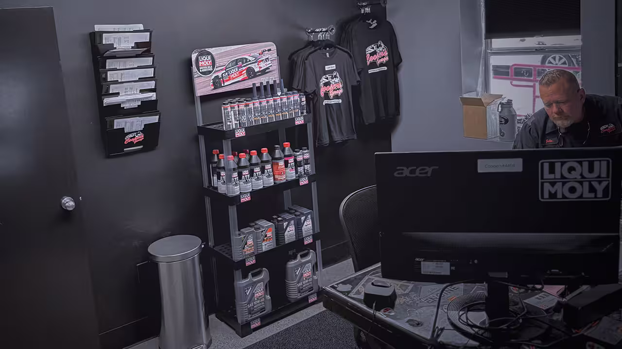 Liqui Moly product display inside Cooper’s Garage office, featuring shelves stocked with additives and oils, branded t-shirts on the wall, and a technician working behind the counter.

