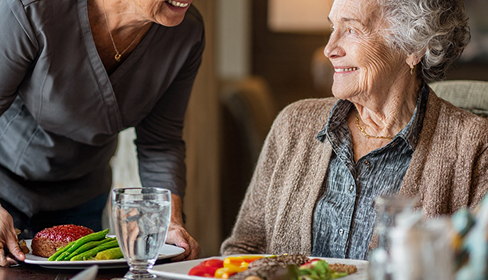 Caregiver serving a plate of food to a smiling elderly woman seated at a dining table.