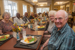 Group of elderly people sitting at a dining table, smiling with plates of food and drinks in front of them in a well-lit room.