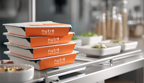 Stack of four orange and white Nutré meal plan boxes on a kitchen counter with blurred bowls of food in the background.