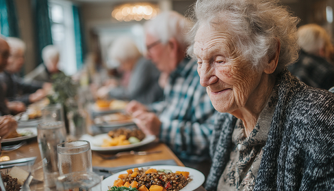 Elderly woman smiling while sitting at a dining table with a plate of food, surrounded by other seniors in a communal setting.