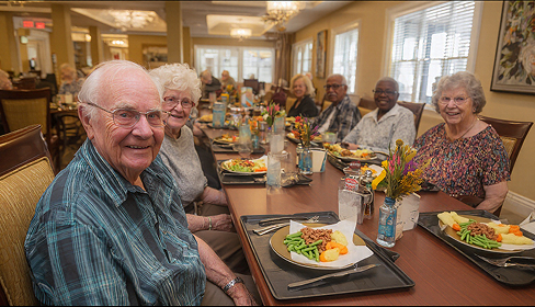 Group of smiling elderly people seated around a dining table with plates of food in a well-lit room.