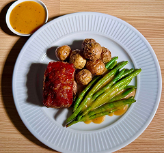 Plate with glazed grilled green beans, roasted baby potatoes, and a piece of barbecued meat with a cup of sauce on the side.