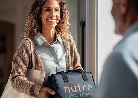 Smiling woman handing over a black insulated bag with Nutre Meal Plans logo through a doorway.