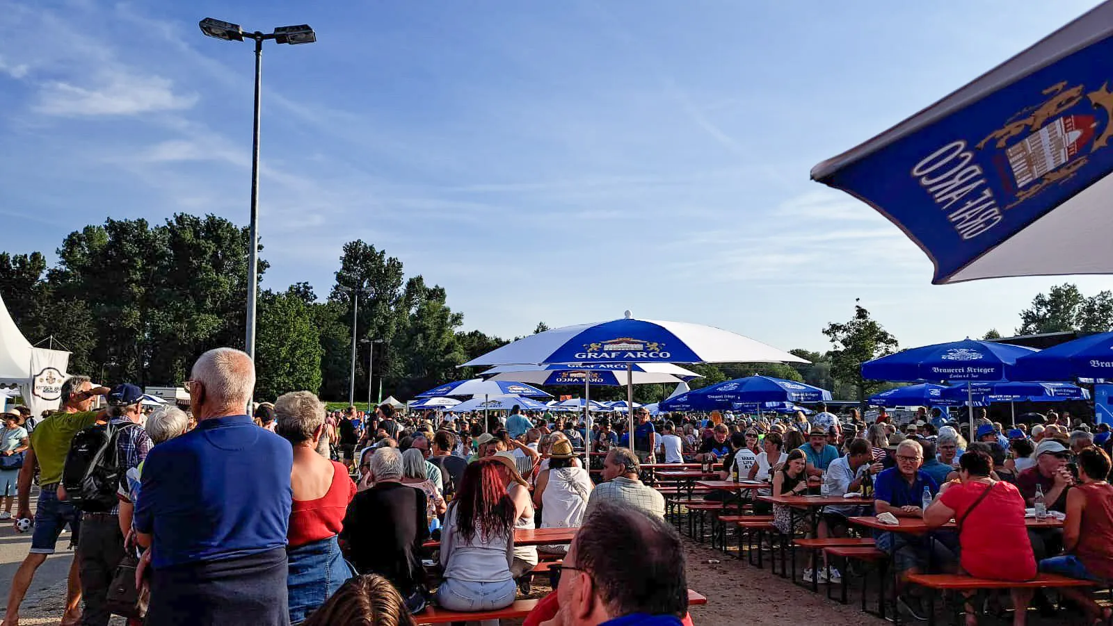 An einem sonnigen Tag sitzt eine große Menschenmenge auf roten Bänken und Tischen unter blau-weißen Sonnenschirmen auf einem Freiluftfest oder in einem Biergarten, mit Bäumen und klarem Himmel im Hintergrund.