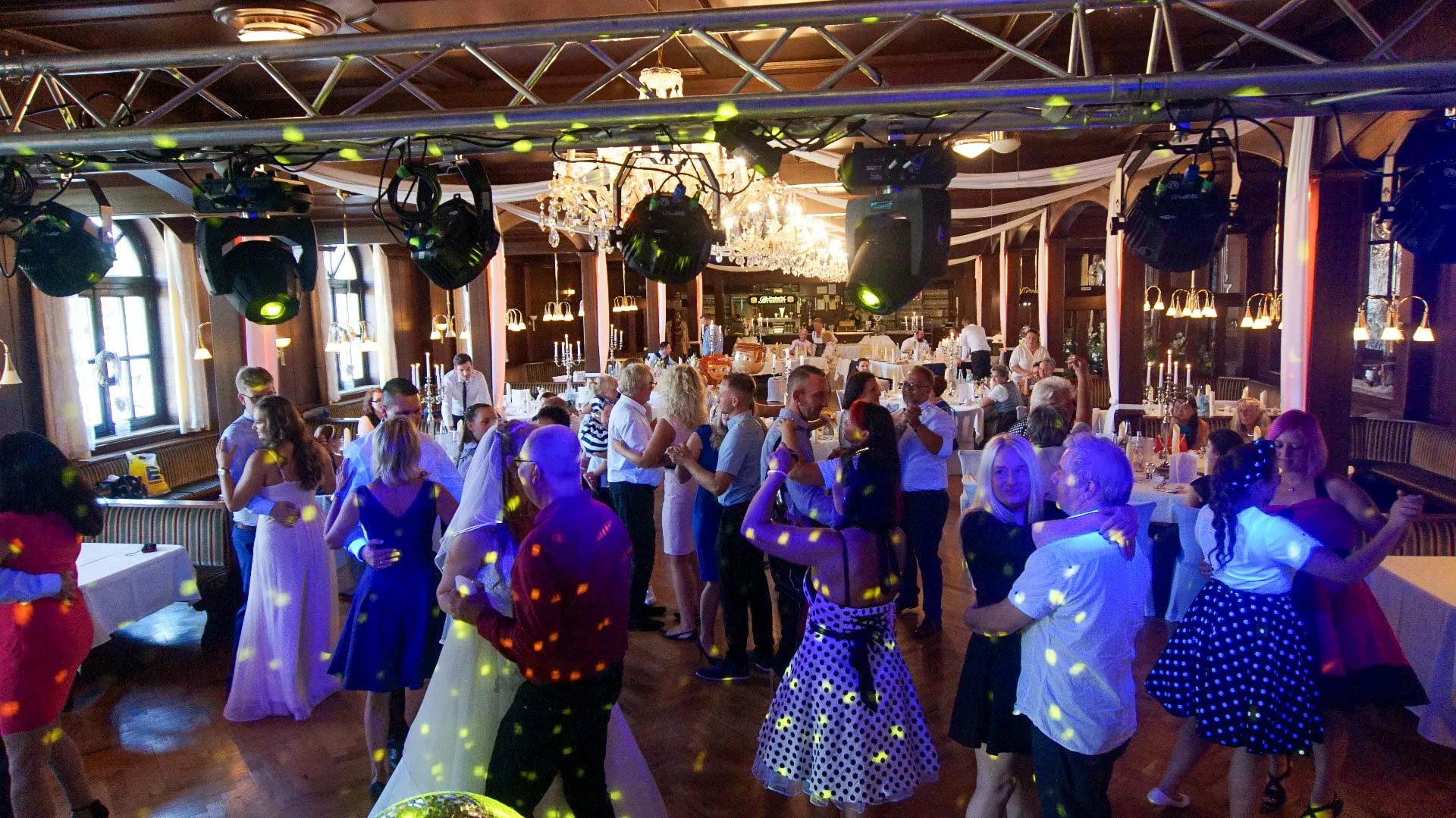 Bei einer festlichen Veranstaltung in einer Halle tanzen Menschen in halb-formellen und gepunkteten Retro-Outfits paarweise miteinander. Bunte Lichter und Kronleuchter erhellen den Raum, der mit dekorierten Tischen und Gästen gefüllt ist.