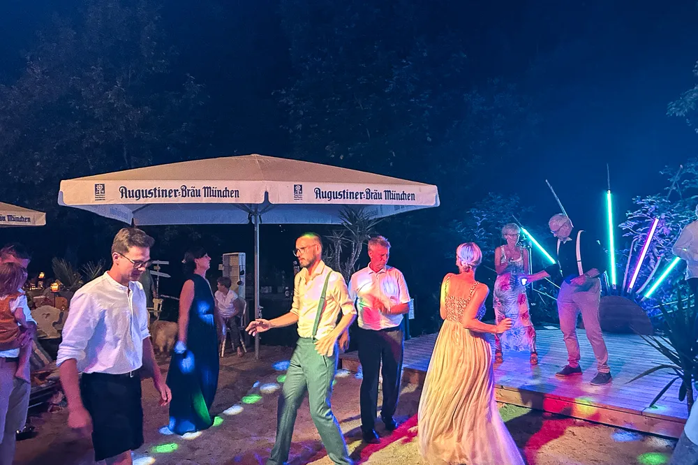 Feierlich gekleidete Menschen tanzen nachts im Freien unter einem Regenschirm von Augustiner-Bräu München, mit bunten Lichtern und einem DJ auf einer kleinen Bühne im Hintergrund. Bäume und eine festliche Atmosphäre umgeben die Szene.