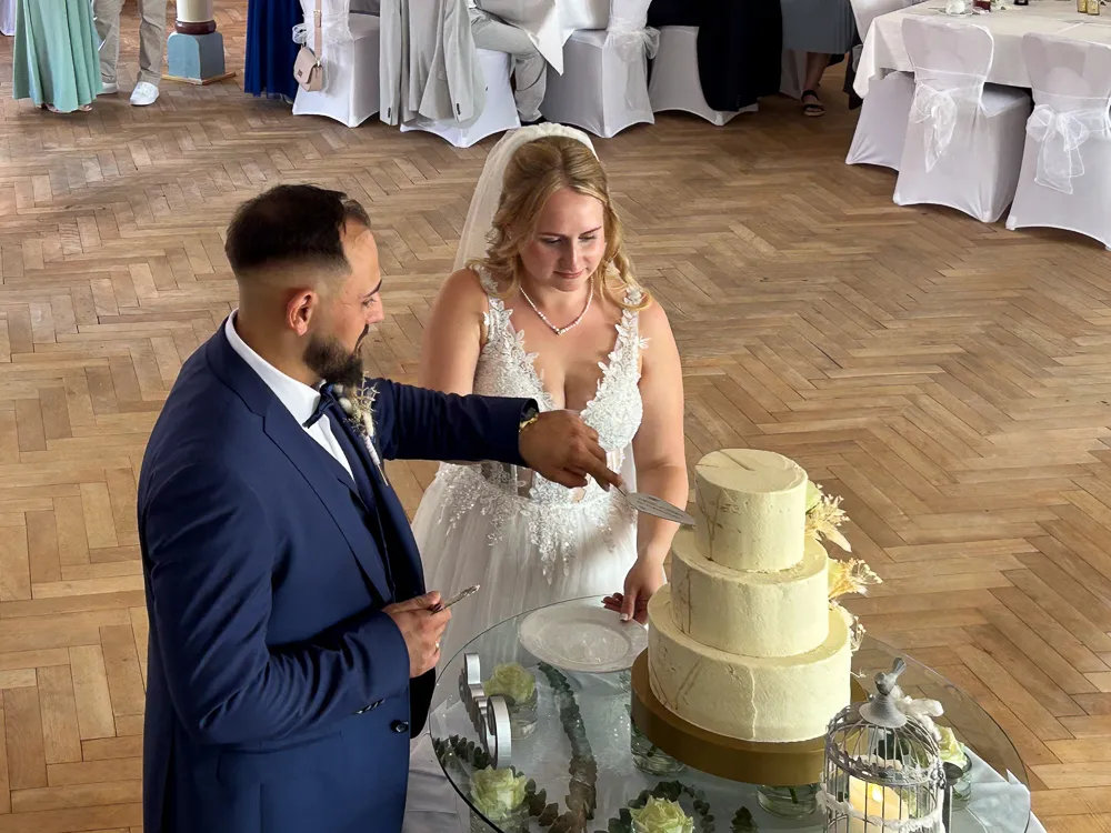 Eine Braut und ein Bräutigam in Hochzeitskleidung schneiden bei einem Empfang gemeinsam eine dreistöckige weiße Torte an. Die Torte steht auf einem Glastisch mit Blumenschmuck in einem Saal mit Parkettboden.