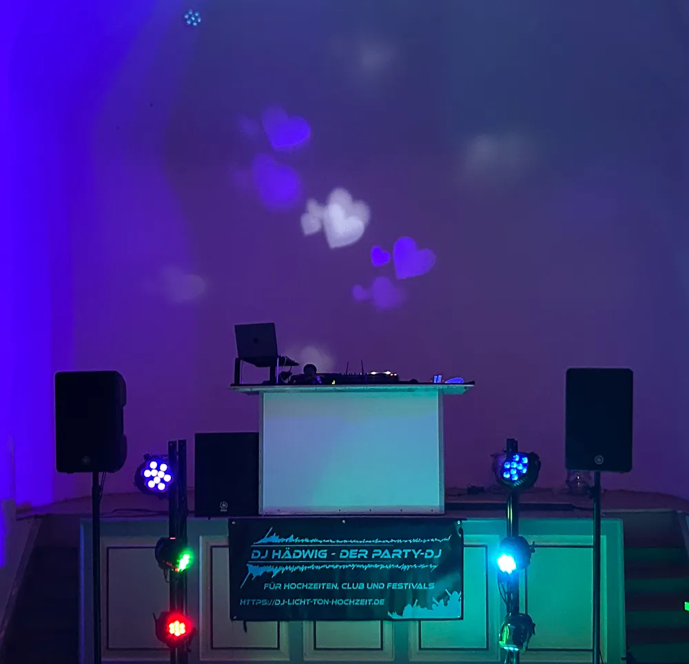 Ein DJ-Pult mit bunten Lichtern und herzförmigen Projektionen an einer Wand, flankiert von Lautsprechern und Beleuchtungsanlagen, aufgebaut für eine Party oder Veranstaltung.