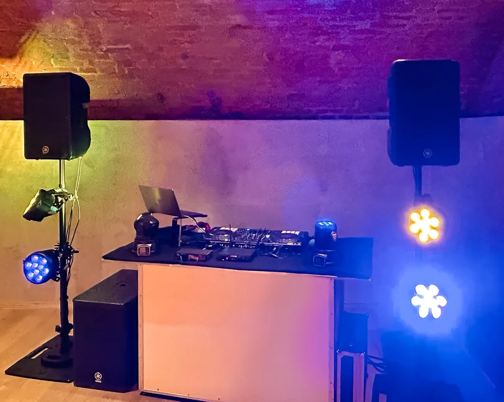 Ein DJ-Setup in einem Raum mit Backsteinwänden, einschließlich Lautsprechern, buntem Bühnenlicht, einem Laptop und DJ-Equipment auf einem Tisch. Die Lichter werfen blaues, gelbes und grünes Licht und schaffen eine Partyatmosphäre.