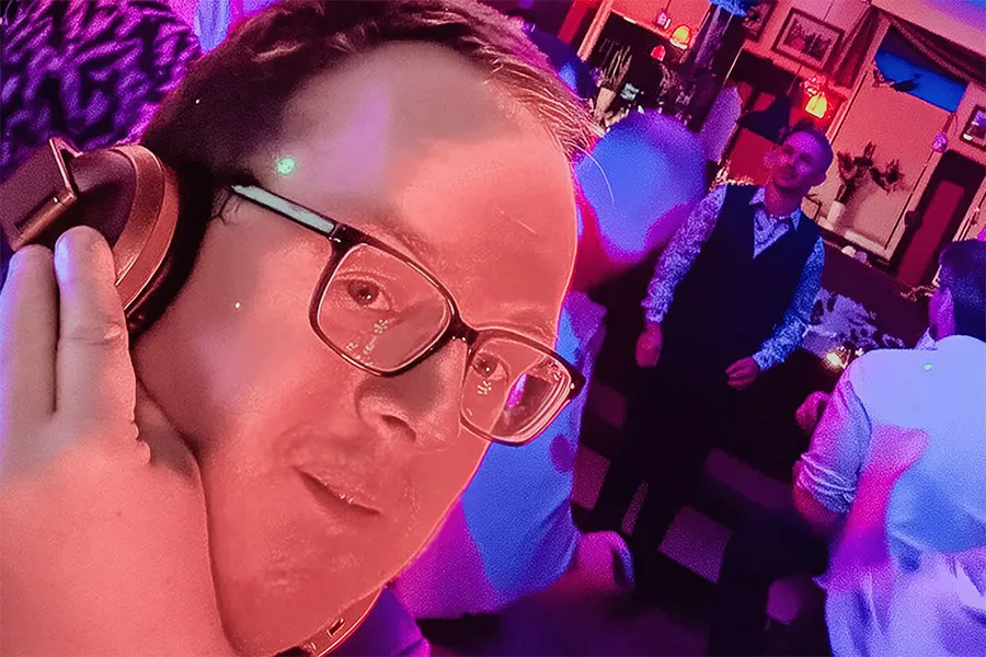 Im Vordergrund ist ein DJ mit Brille und Kopfhörern zu sehen, der von rosa und lila Lichtern angestrahlt wird, während im Hintergrund Menschen tanzen und sich in einer lebhaften, farbenfrohen Partykulisse unterhalten.
