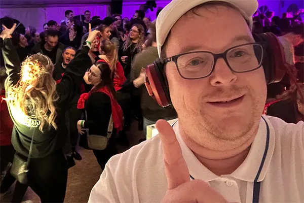 Ein Mann, der eine Brille, Kopfhörer und eine weiße Mütze trägt, lächelt und zeigt nach oben, während er ein Selfie auf einer lebhaften Indoor-Party macht, auf der die Leute im Hintergrund tanzen und sich amüsieren.