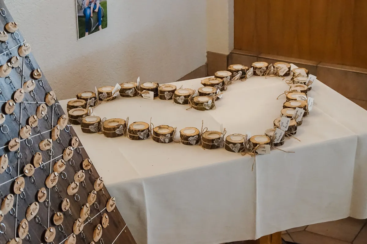 Tisch mit einer weißen Tischdecke, auf dem kleine Holzkerzenhalter in Herzform arrangiert sind, jeder mit einem Namensschild und Juteband, daneben ein Brett mit vielen hängenden ovalen Holzanhängern mit Namen.