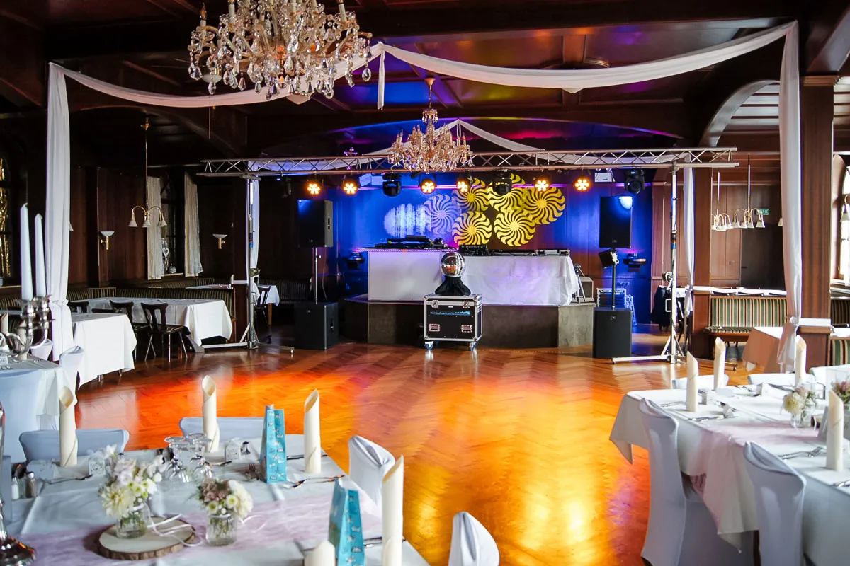 Eleganter Festsaal mit Holzboden, geschmückten Tischen mit weißen Tischdecken und Kerzen, Kronleuchtern an der Decke, einer DJ-Bühne mit Beleuchtung und Lautsprechern im Hintergrund.