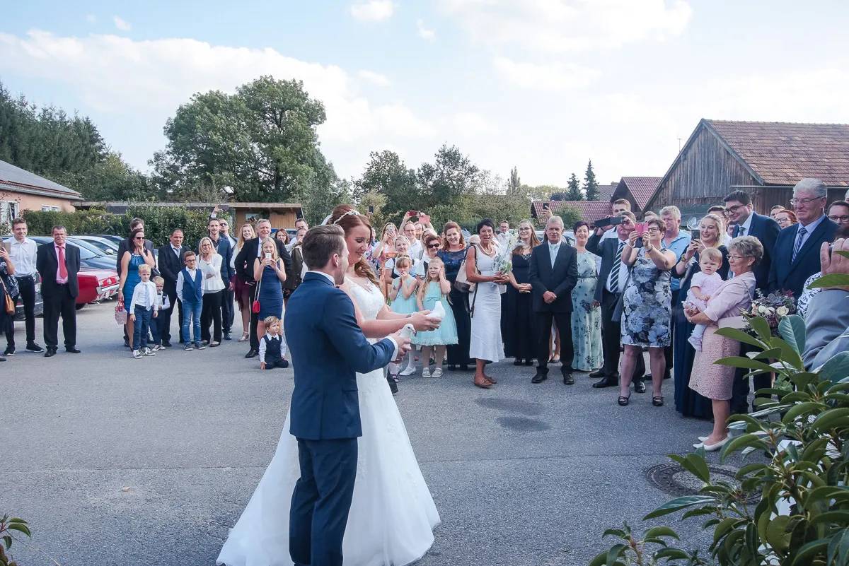 Braut und Bräutigam in formeller Kleidung stehen zusammen im Freien, umgeben von vielen Gästen, die Fotos machen und zuschauen, unter einem bewölkten Himmel mit Bäumen und Gebäuden im Hintergrund.