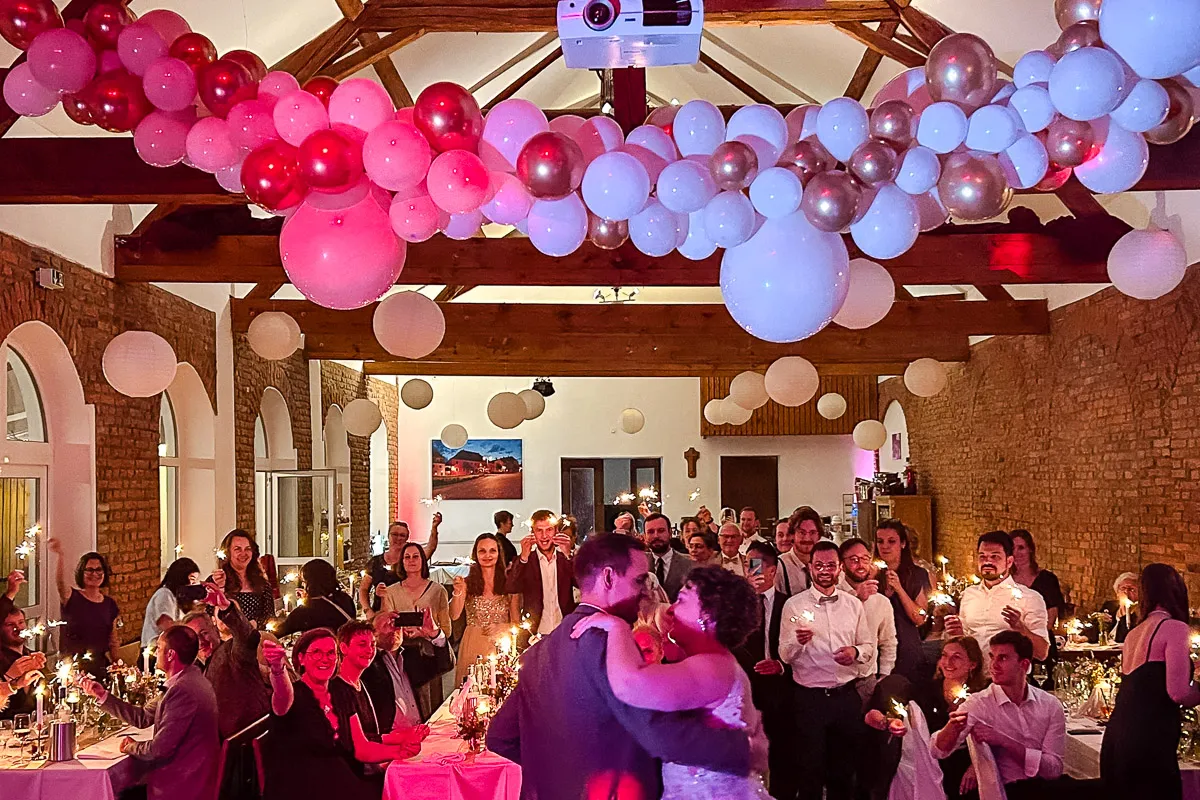 Ein Hochzeitsfeier mit vielen Gästen, die einen Tanzsaal mit weißen, rosa und roten Luftballons an der Decke schmücken, während ein Paar in der Mitte tanzt und die Gäste funkelnde Wunderkerzen halten.