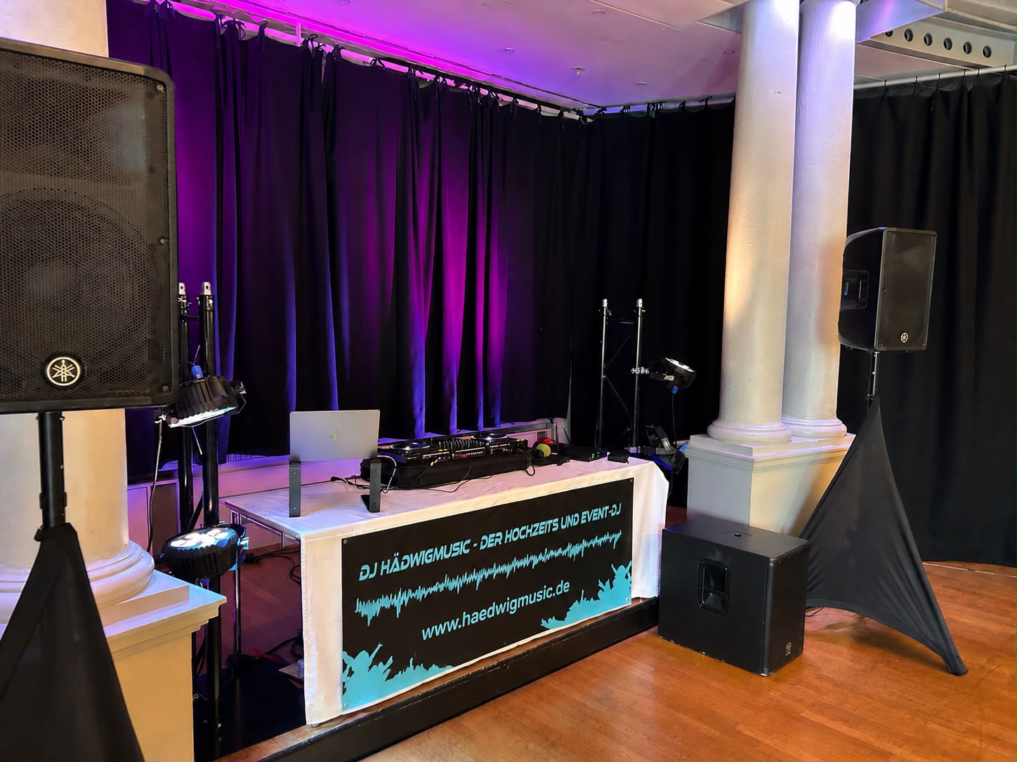 DJ-Setup auf einer Bühne mit zwei weißen Säulen, schwarzem Vorhang und lila Beleuchtung, Lautsprecher und DJ-Equipment auf einem weißen Tisch mit einem Banner.