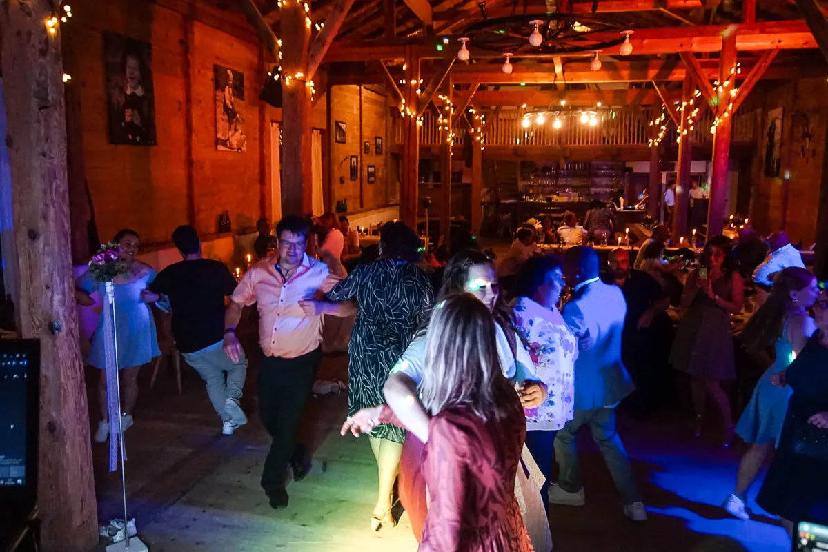 Menschen tanzen und feiern in einem rustikalen, holzverkleideten Veranstaltungsraum mit warmen Lichtern und Girlanden, die eine festliche Atmosphäre schaffen.