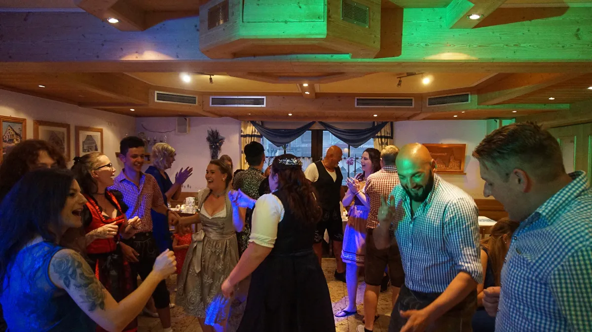 Menschen feiern fröhlich in einem Raum mit Holzdecke und bayerischer Tracht.