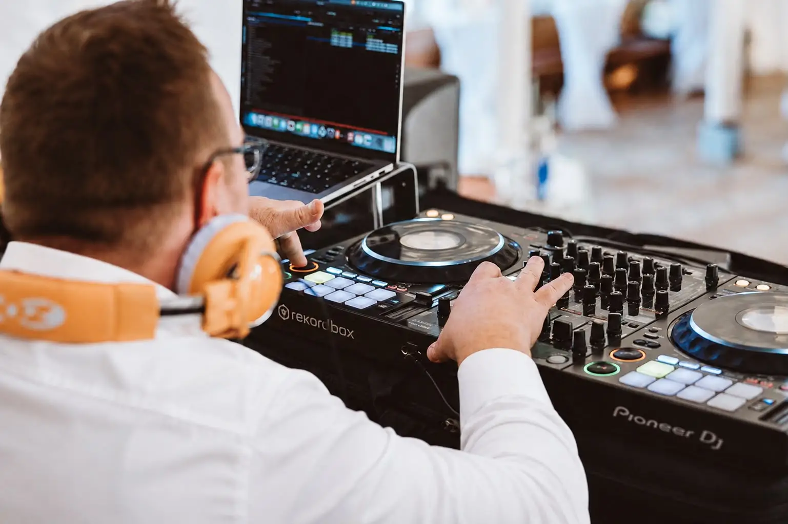 DJ mit orangen Kopfhörern bedient Pioneer DJ Mischpult und Laptop bei Veranstaltung.