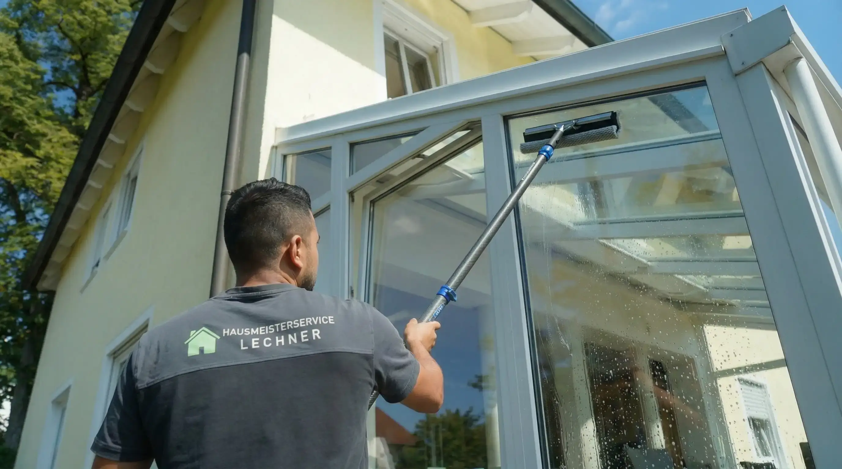 Mann wäscht mit einem Fensterreiniger ein großes Glasfenster an einem Haus.